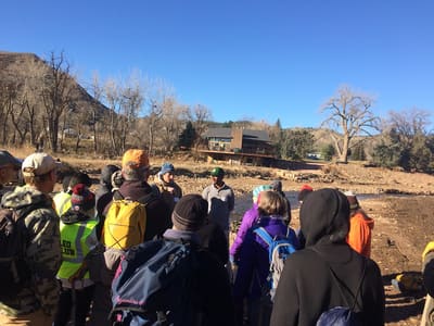 A crowd of volunteers in hats and jackets gathered on the riverbank for a replanting day, cottonwoods and cliffs behind